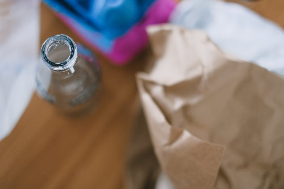 A clear glass bottle with a narrow neck and threaded rim, positioned on a wooden surface, with the bottle's opening facing upward. The bottle appears to be empty or contains a small amount of liquid, and it has a smooth, transparent texture reflecting light subtly. In the background, partially out of focus, is a colorful carton or container with hues of blue, pink, and purple, along with crumpled brown paper or packaging material, which is loosely arranged and slightly wrinkled. The setting suggests an indoor environment, possibly a kitchen or a workspace, where waste or recyclables such as glass containers and packaging materials are being collected or prepared for disposal or recycling. Rubbish Removal Waterloo’s involvement in local waste management may relate to the collection or removal of such materials, emphasizing environmentally responsible waste handling practices. The scene is lit naturally or with soft indoor lighting, highlighting the textures and colors while maintaining a neutral, professional tone suitable for a rubbish removal service context.
