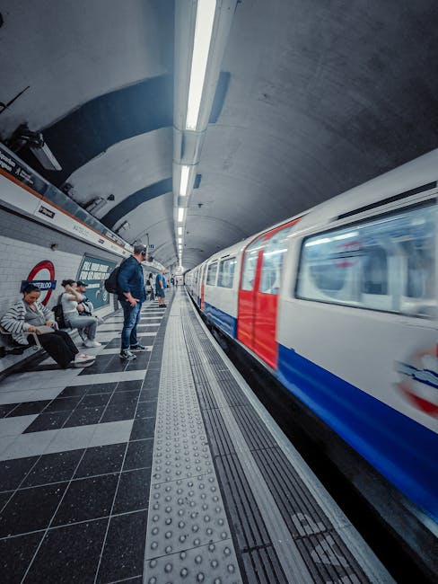 Inside Waterloo underground station, the platform is illuminated by bright, evenly spaced fluorescent lighting fixtures attached to the curved concrete ceiling. Several passengers are waiting on the left side of the platform, sitting on black benches with silver metal supports; they are dressed in casual clothing, some with backpacks and luggage, indicating commuter activity. A man in a dark jacket stands near the edge of the platform, looking towards the approaching train. The train itself is moving quickly, with a streamlined white body accented by red and blue horizontal stripes running along its side, creating a slight blur effect due to motion. The platform has a tactile paving strip with raised bumps near the edge for safety, and the flooring consists of black and dark gray tiles arranged in a checkered pattern, some glazed and others matte. The background shows part of the station’s tiled white walls with station signage, emphasizing the urban underground environment. This scene reflects typical private or independent collection practices for urban rubbish removal, where efficient station access and passenger flow are crucial, indirectly supporting the need for professional waste handling in busy transit hubs. Rubbish Removal Waterloo specializes in professional waste management services, including rubbish clearing from commercial and residential sites, often requiring on-site clearance solutions in high-traffic areas such as this.
